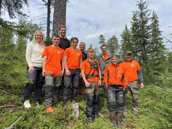 Gruppebilde av ni personer som står i en skog. På første rad, i oransje klær, står ungdommene som har sommerjobb.